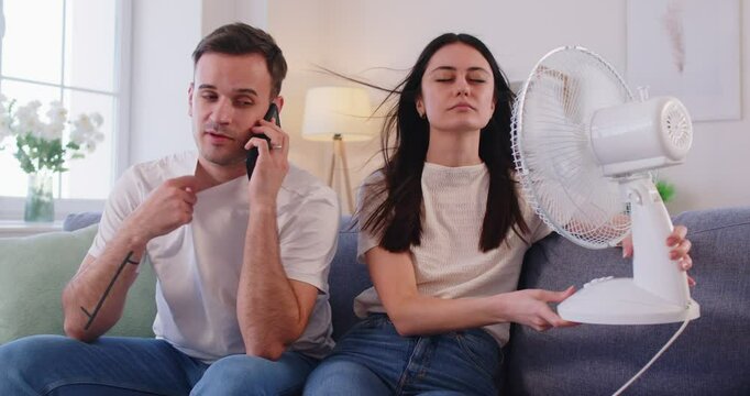Family couple suffering from summer heat, feeling hot, using electric fan, struggling with lack of air condition, posing on sofa in living room, getting cool or refreshed. Overheating