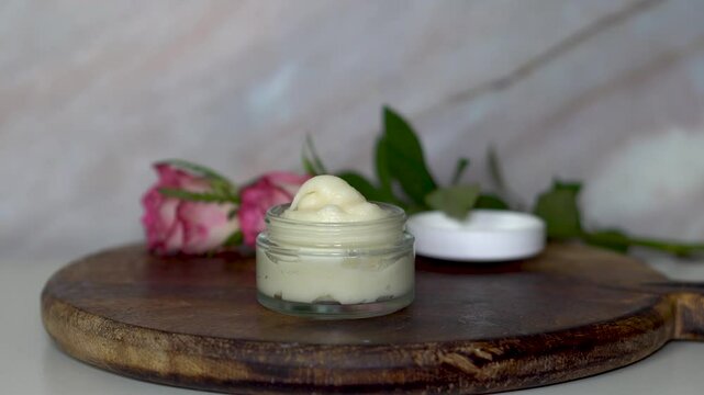 Homemade natural cream in clear glass jar on rustic dark wooden board, decorated with fresh pink roses. Organic skincare product, perfect for beauty, spa, wellness, and cosmetic themes.
