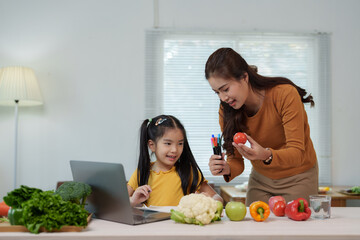 Asian mother showing tomato to her daughter and teaching her online during homeschooling, with various fresh vegetables and fruits on table, promoting healthy eating habits for children