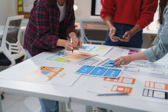 Web designers collaborating on user interface and user experience design for a mobile app, discussing and arranging wireframes on a desk while brainstorming innovative ideas together