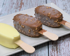 Delicious scene with three ice cream bars, including chocolate and vanilla flavors, served on a white plate with toppings, seeds, nuts, and colorful sprinkles. Rustic wooden background