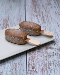 Delicious scene with three ice cream bars, including chocolate and vanilla flavors, served on a white plate with toppings, seeds, nuts, and colorful sprinkles. Rustic wooden background 