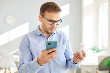 Concerned young man in glasses holding smartphone and medicine bottle in bright room. Worried...