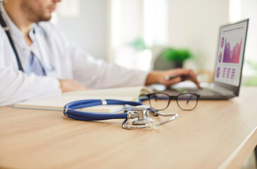 Stethoscope lying on the table near a doctor or nurse working online with a laptop in a hospital or...