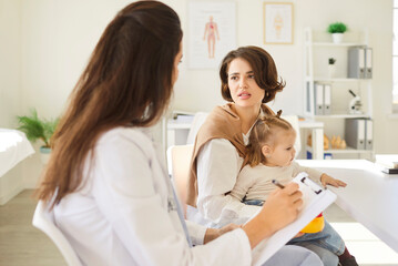 Pediatrician doctor treats newborn during visit in hospital, making notes, mother holding baby girl, little patient at consultation in clinic, small child healthcare for regular health examination
