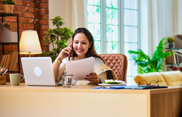 Speaking on mobile, Indian lady in saree reads business documents at modern office workspace