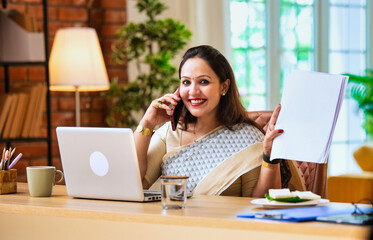 Speaking on mobile, Indian lady in saree reads business documents at modern office workspace