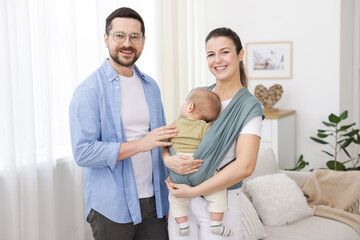 Fototapeta premium Portrait of happy family indoors. Mother holding her child in sling (baby carrier) at home