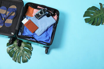 Open suitcase with different clothes, camera and passport on light blue background, above view....