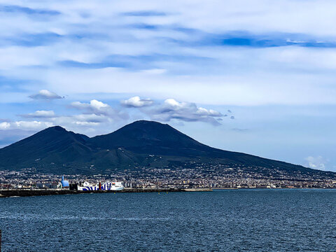 A massive mountain rises from the deep blue sea under a sky with scattered clouds. The rugged peak contrasts with the calm water, capturing a dramatic coastal scene., vesuvio, naples, napoli, napoles