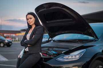 Young woman leaning on broken car with open hood, standing outdoors. Waiting for help. Car service,...