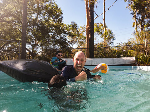 Man in his fifties playing with kids and grandchild in backyard pool in summer laughing out loud