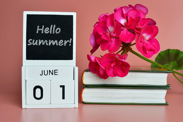 Hello summer text on a black chalkboard. A wooden calendar stands next to books and a flower.