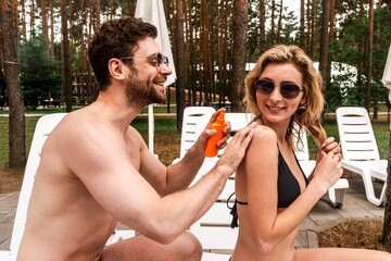 Smiling guy seated on the lounger squeezing a sunscreen lotion out of the plastic bottle to the...