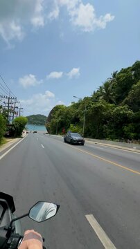 A first-person perspective from a motorcycle rider traveling along a winding coastal road. A large, natural rock formation dominates the right side of the frame, bordered by lush green vegetation. In