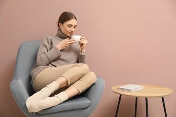 Beautiful woman with cup of drink relaxing in comfortable armchair near beige wall indoors