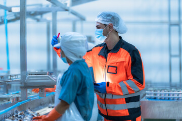 Supervisor in orange uniform and hairnet inspects canned food on the production line with factory...