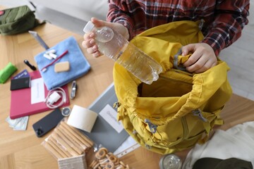 Survival kit. Woman putting bottle of water into backpack at wooden table indoors, above view