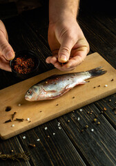 A person is seasoning a whole fish with spices while it sits on a wooden cutting board. The kitchen setting has a warm, natural atmosphere, enhancing the culinary process
