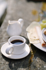 Pretty small white cup with the black coffee on the wooden table with ceramic milk pot and plate full of cheese and fruits i the mmorning garden