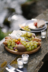 The wooden plate full of fresh fruits, vegetables, bread and cheese on the table in the mountains near the river. Lovely  picnic in the forest in the sunny spring day 