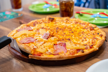 Freshly baked Hawaiian pizza topped with melted cheese, ham, and pineapple, served on a wooden plate with drinks and colorful tableware in the background