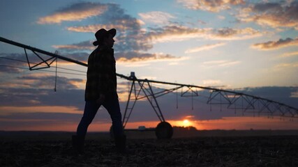 Farmer walks through field at sunset. Large machinery prepares soil for planting. Farmer observes farming process. Rural agriculture lifestyle. Farmer field machinery soil preparation concept.