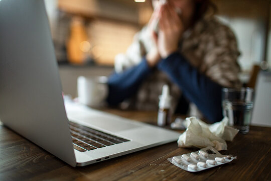 Sick woman sneezing while working from home with laptop in kitchen