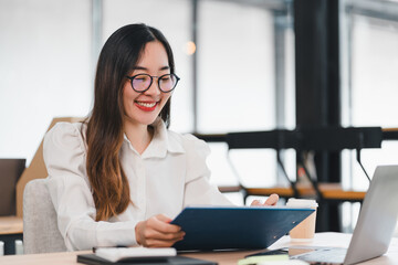 Young woman smiling while reading document in modern office with laptop and notebook
