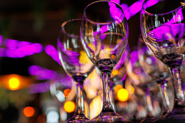 Empty clear wineglasses stand in rows on bar counter at nightclub, blurred purple neon and yellow lights on background. Clean cocktail glasses ready for celebrate party.
