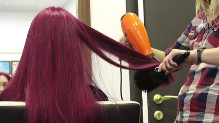 Hairstylist using orange hair dryer and round brush styling vibrant burgundy red hair in salon indoor. Concept of hairdressing, beauty salon, hair care service, color maintenance. - Powered by Adobe