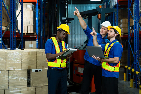 Three warehouse workers wearing safety vests and helmets working together checking inventory using digital tablet and laptop teamwork concept efficiency logistics industry diversity collaboration