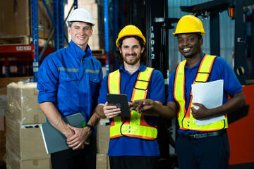 Three diverse warehouse workers wearing helmets and reflective safety vests smiling holding tablet...