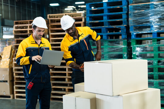 Two warehouse workers wearing yellow uniforms and white helmets working together scanning and checking packages using laptop representing teamwork logistics efficiency diversity industrial workplace