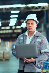 Warehouse male worker wearing white helmet standing firmly holding laptop ready for logistics...