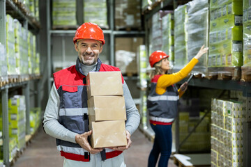 Portrait of a warehouse worker holding boxes while a female worker is taking inventory of goods in the warehouse in the background