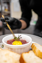 A chef garnishes a dish of steak tartare topped with a raw egg yolk and fresh herbs, served with slices of bread, showcasing artful presentation in a professional kitchen.
