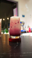 A vibrant iced purple latte topped with creamy foam, served in a tall glass on a black marble table. The cozy and modern cafe background with soft lighting enhances the refreshing and artistic appeara