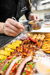 A chef applies a rich glaze to a flavorful assortment of ribs and grilled meats in a takeout box, complemented by corn and sides, showcasing an inviting culinary presentation.