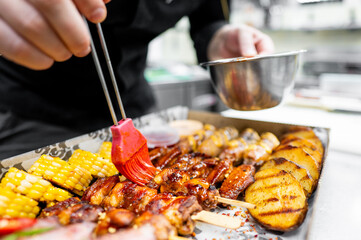 A chef uses a brush to apply a vibrant sauce to a platter of grilled meats, including ribs and skewers, alongside corn and grilled potatoes in a stylish takeout box.