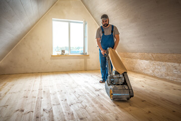 Professional worker carpenter in blue overalls grinding sanding a wooden floor by using floor sander in cockloft. Floor renovation