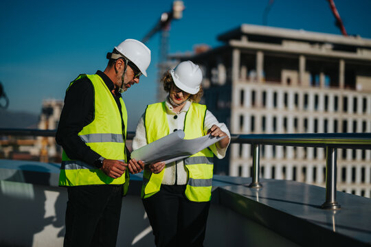 Two architects in safety gear discuss construction plans on-site. They wear reflective vests and helmets while examining blueprints. The background shows an under-construction building structure.