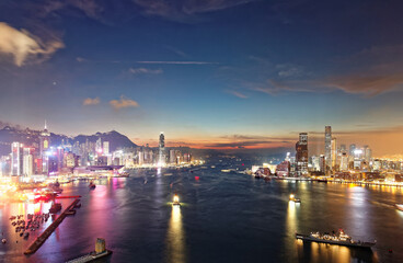Fototapeta premium Scenery of Hong Kong at nightfall, with a city skyline of modern skyscrapers by Victoria Harbour between Kowloon & Hongkong Island and ships navigating in the busy seaport under beautiful twilight sky