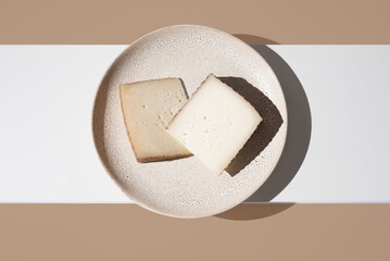 Dos trozos de queso de oveja curado y de cabra en un plato sobre fondo blanco y marrón. Vista superior	