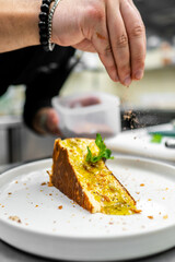 A chef sprinkles a dusting of seasoning over a slice of flavorful cake, garnished with fresh mint, on a sleek white plate, enhancing the dish's presentation in a modern kitchen.