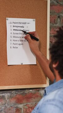 African-American man crosses out accomplished to-do list points on cork board near brick wall closeup. Black worker checks plan of flat renovation