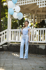 Young woman holding blue and white balloons in a park during a sunny day while wearing a formal light blue outfit