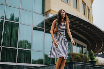 Obraz premium A young woman joyfully laughs outside a modern building. She is wearing a stylish striped dress, exuding happiness and energy. The glass facade adds a contemporary urban feel.