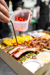 A chef adds fresh chili slices to a vibrant platter of grilled meats and corn, showcasing meticulous garnishing in a takeout box, enhancing the flavor and presentation.