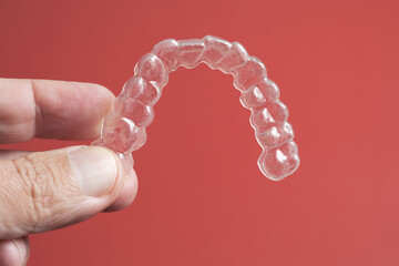 Transparent dental aligner held by a hand against a red background
